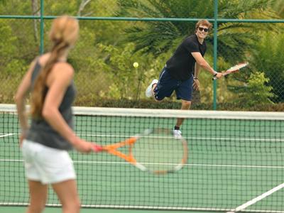 Two people playing tennis on an outdoor court with green fence and trees in the background.