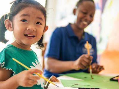 Ein Mädchen malt bunte Bilder, im Hintergrund sitzt eine Frau, beide lächeln.