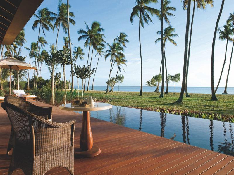 Terrasse mit Holzboden, Sesseln, Pool und Blick auf Palmen und das Meer im Hintergrund.