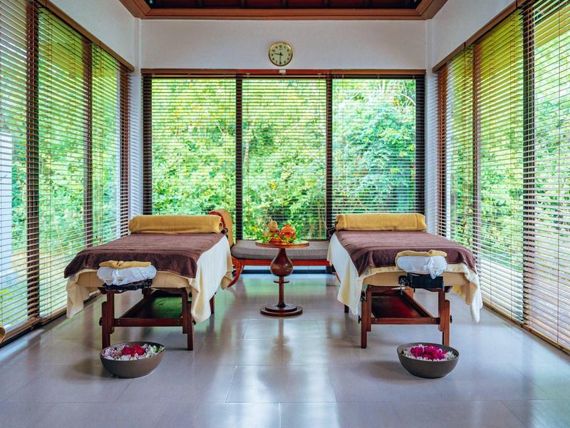 Hell eingerichteter Wellnessraum mit zwei Massageliegen, großen Fenstern und grünem Gartenausblick.