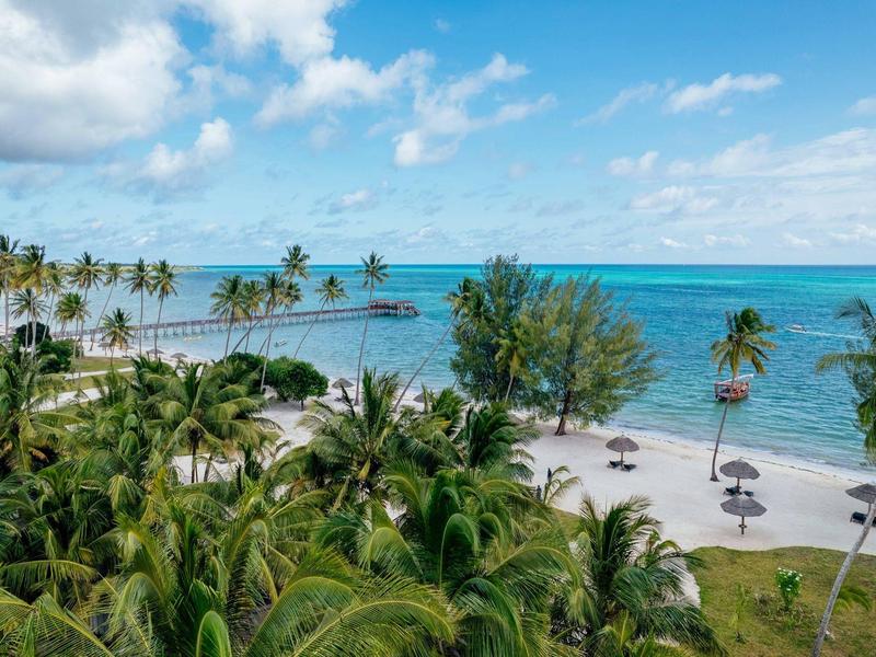 Strand mit weißen Sand, Palmen, Sonnenschirmen und blauem Meer unter hellem Himmel.