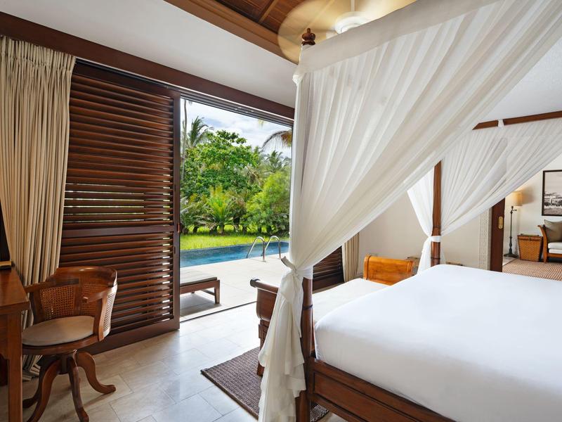 Helles Schlafzimmer mit Himmelbett, Holzmöbeln, Vorhängen und Blick auf einen Pool und Garten.