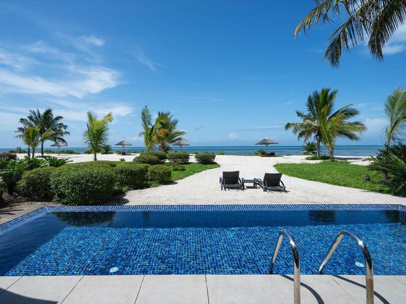 Blick auf blauen Pool, Palmen und weißen Sandstrand unter blauem Himmel mit wenigen Wolken.