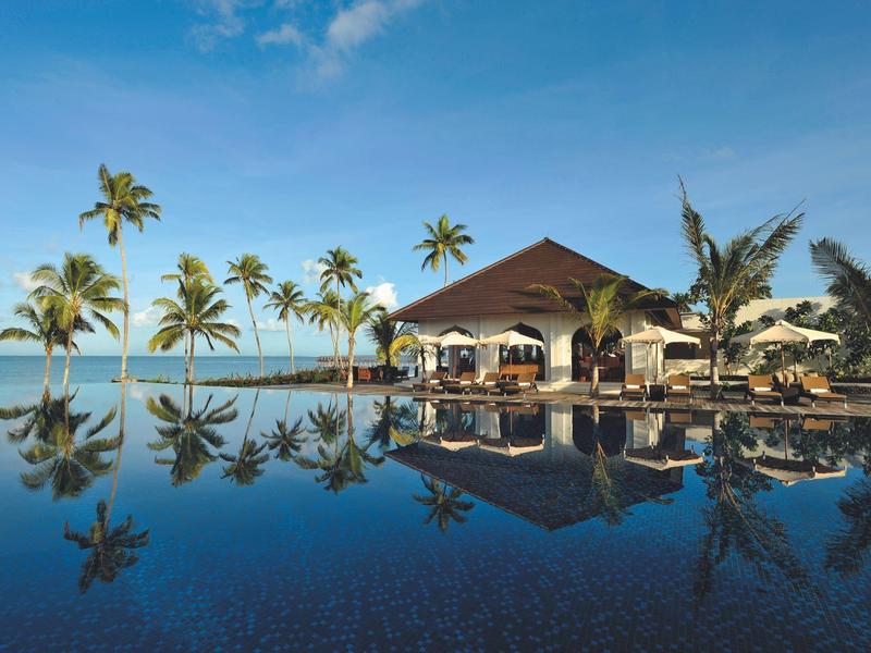 Bungalow am Strand mit Palme und großem ruhigem Pool, der die Szenerie spiegelt.