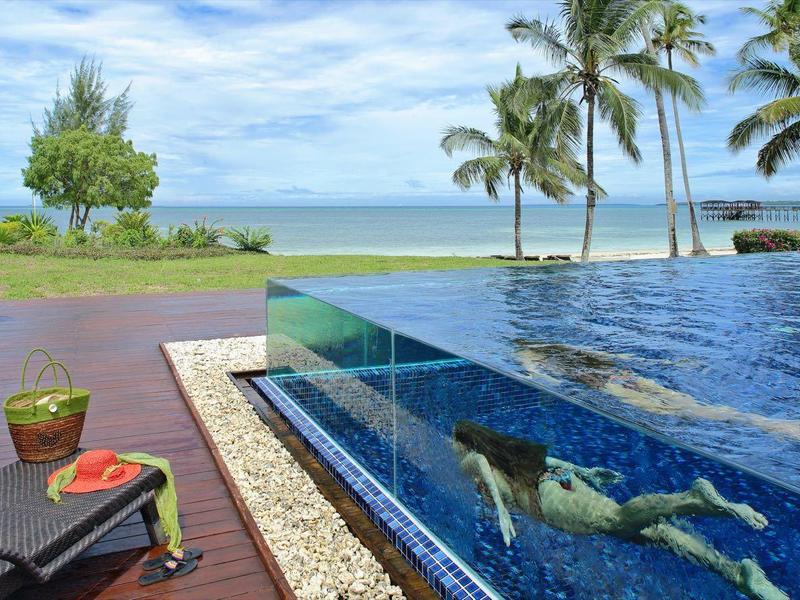 Luxuriöser Pool mit Meerblick, Palmen, grünem Rasen und gemütlichen Sitzplätzen, sonniger Himmel.