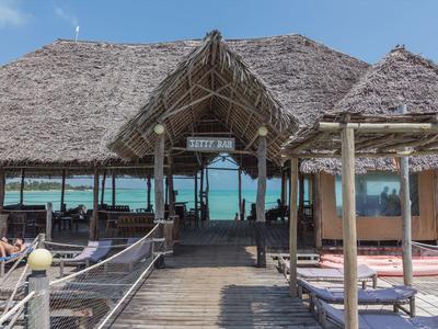 Holzhütte mit Strohdach auf Steg über türkisfarbenem Meer, mit Sitzbereichen und Sonnenliegen.