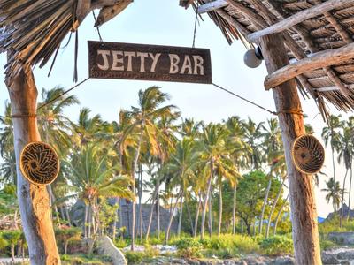 Holzrahmen mit Palmwedel-Dach und Schild "Jetty Bar" vor Palmen und Strand im Hintergrund.