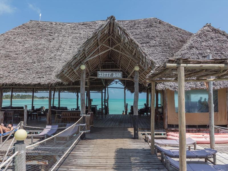 Holzhütte mit Strohdach auf Steg über türkisfarbenem Meer, mit Sitzbereichen und Sonnenliegen.