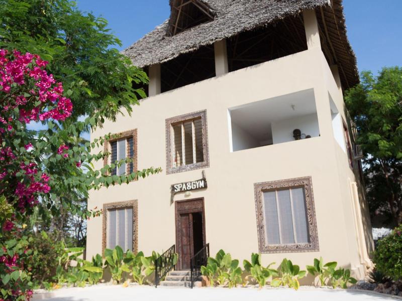 Dreistöckiges Haus mit strohgedecktem Dach, weißen Wänden, Pflanzen und blühenden Sträuchern.