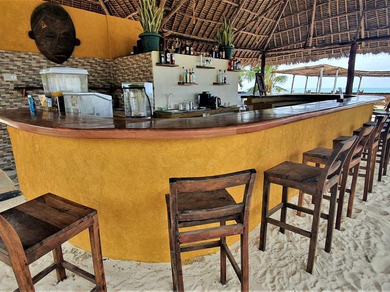 Freiluft-Bar mit gelbem Tresen und Holzstühlen auf Sandboden unter Strohdach.