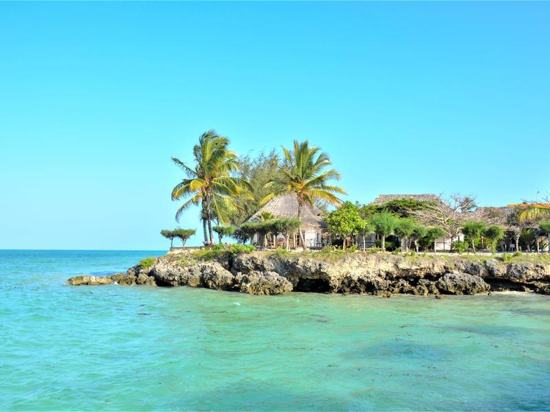 Klares türkisblaues Meer mit felsiger Küste, Palmen und einem kleinen Haus unter blauem Himmel.