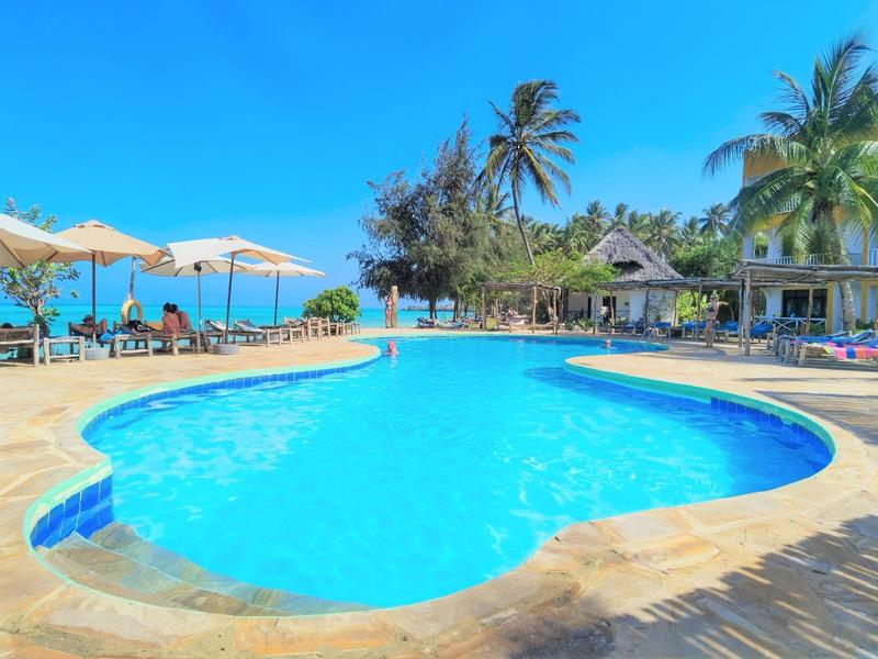 Großer, klarer Pool umgeben von Liegestühlen, Schirmen, Palmen und einem blauen Himmel.