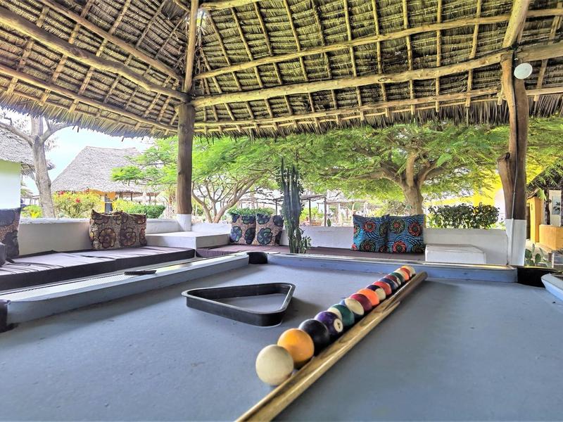 Billardtisch mit einer bunten Kugelgruppe unter einem offenen Strohdach mit Sofas und grüner Umgebung.