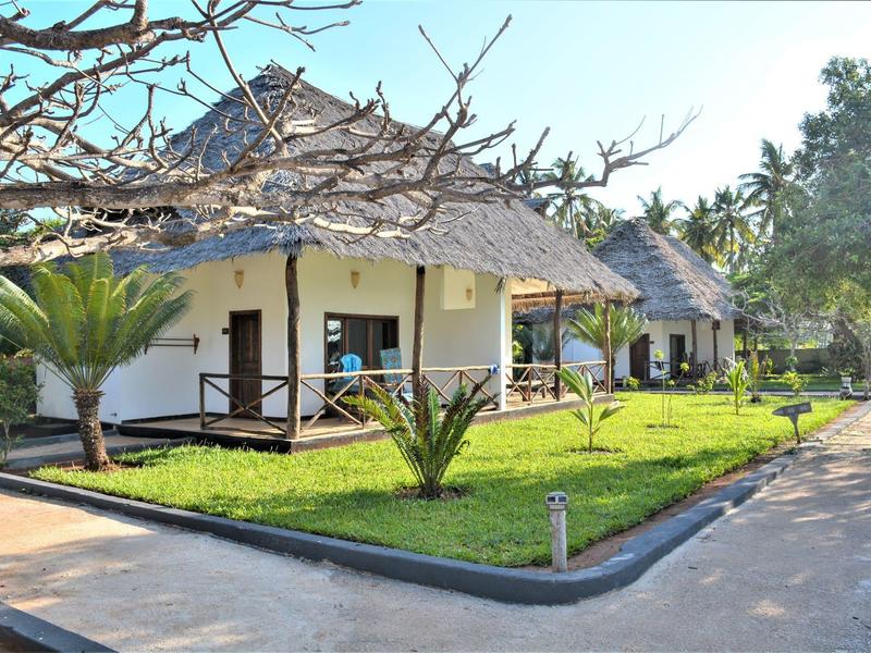 Weiße Bungalows mit Strohdächern, grüner Rasen und Palmen, sonniger Himmel im Hintergrund.
