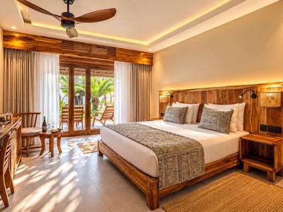 Chambre d'hôtel lumineuse avec grand lit, mobilier en bois et balcon avec plantes.