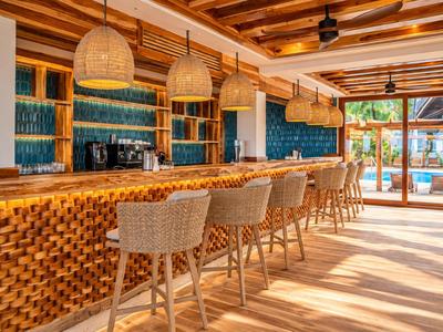 Espace bar d'hôtel moderne avec comptoir en bois, tabourets tissés et carreaux bleu-vert.