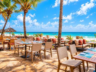 Restaurant de plage ouvert avec tables en bois, chaises, palmiers sous ciel bleu et mer.