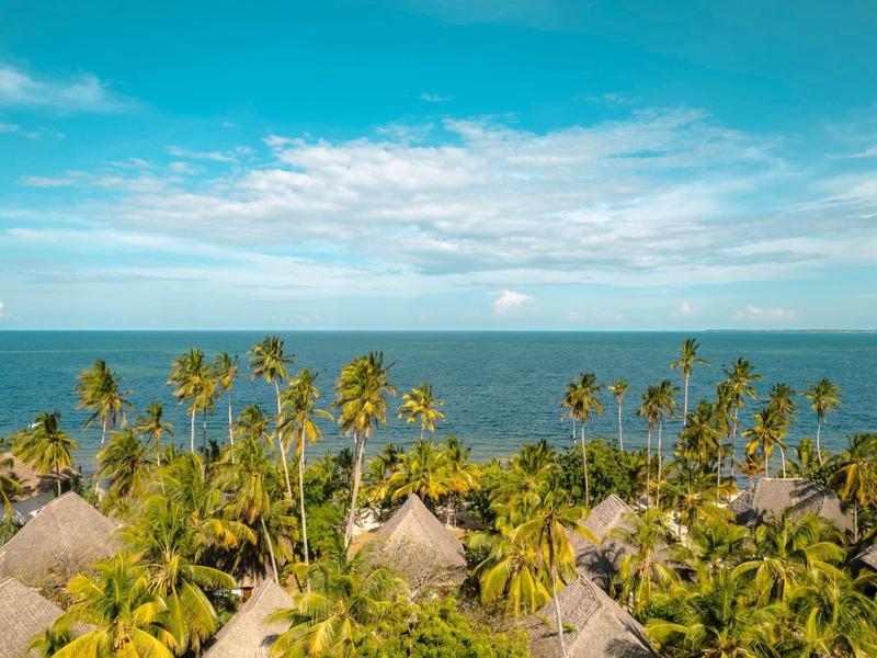 Tropische Küste mit Palmen, Strohdachhäusern und blauem Himmel über ruhigem Ozean.