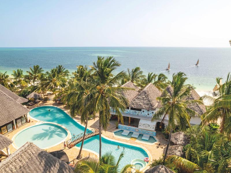 Hotel mit Poolbereich zwischen Palmen am Strand mit Blick auf das Meer.