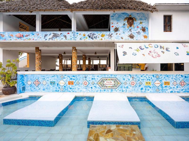 Moderner Poolbereich mit blauer Mosaikfliesenwand, weißen Liegen und offenem Gebäude im Hintergrund.