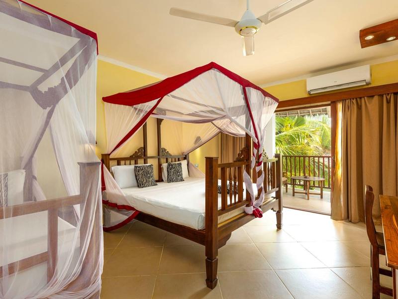 Hell eingerichtetes Schlafzimmer mit Himmelbett, Moskitonetz, Ventilator und Balkon mit Blick ins Grüne.