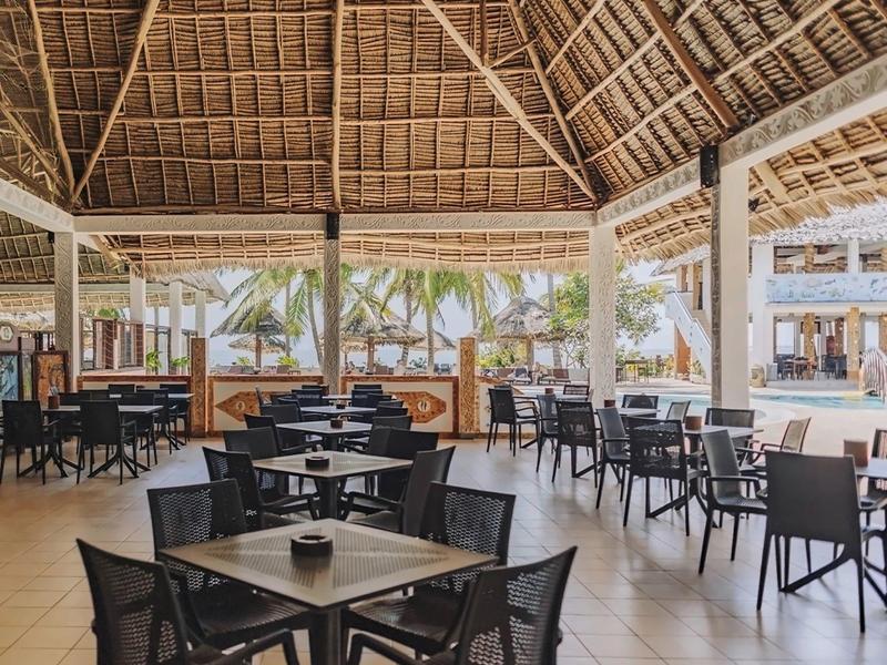 Offener, überdachter Essbereich mit Holztischen, schwarzen Stühlen und Blick auf Palmen draußen.