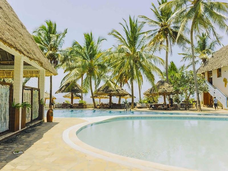 Ein Pool umgeben von Palmen und Gebäuden unter klarem Himmel an einem tropischen Strand.