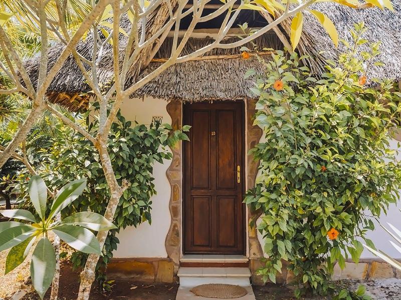 Ein hölzerner Eingang mit einer geschlossenen Tür, umgeben von grünen Pflanzen und einem Strohdach.