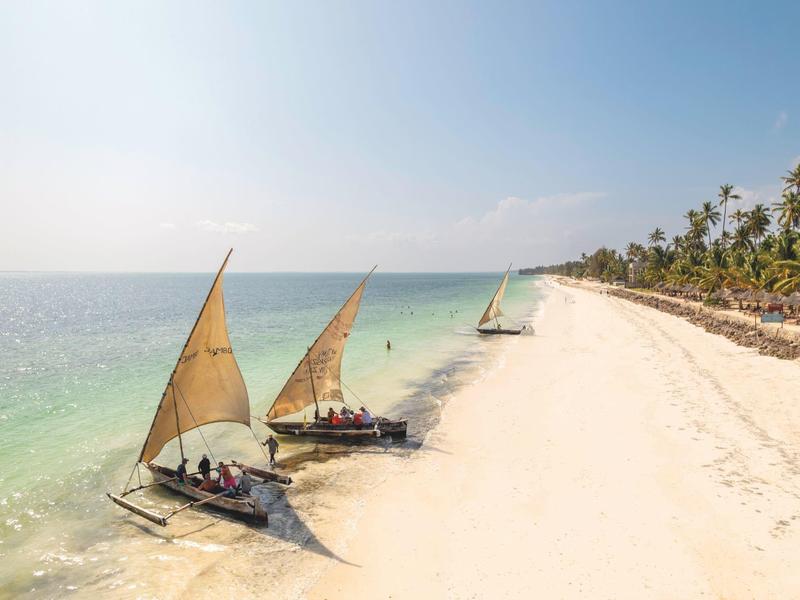 Drei Segelboote segeln nah am weißen Sandstrand mit Palmen unter klarem Himmel.