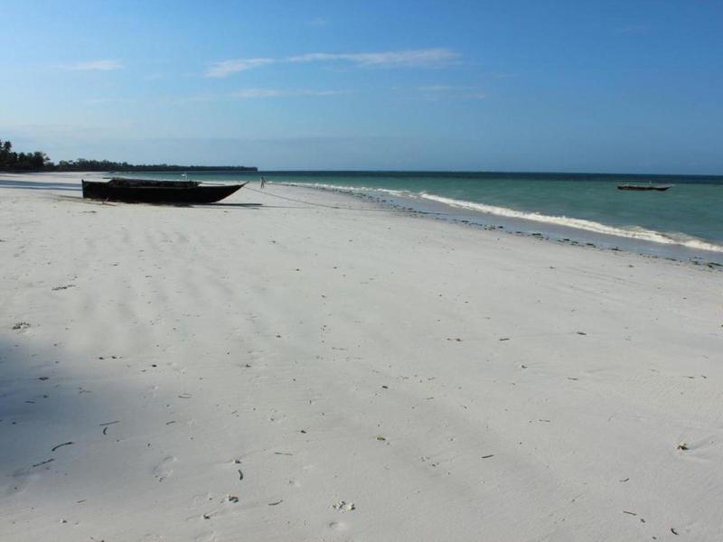 Weißer Sandstrand mit ruhigem Meer und blauem Himmel, links einige Palmen und Holzboote.