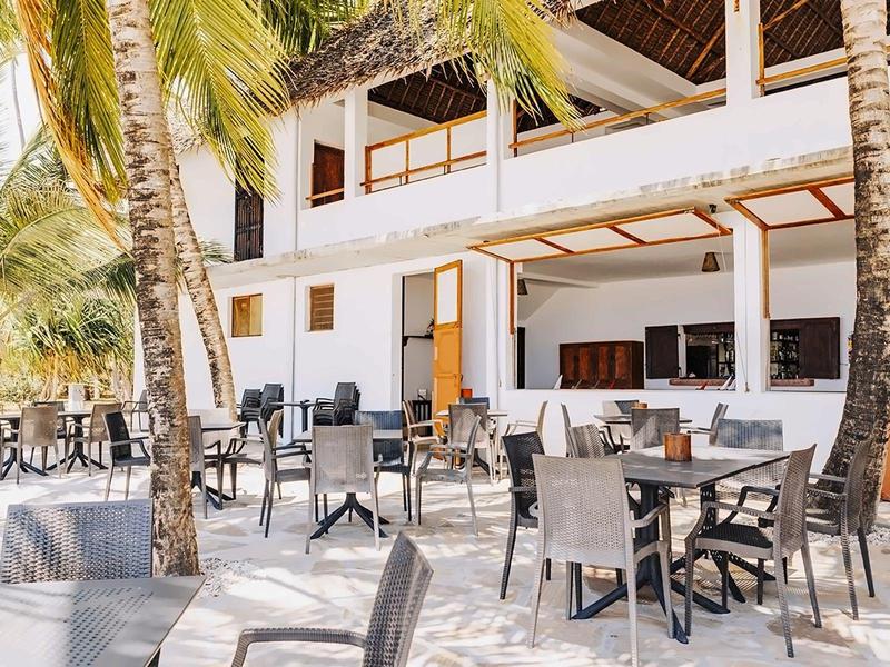 Offenes Restaurant mit schwarzen Tischen und Stühlen, Palmen und weißer zweistöckiger Fassade.