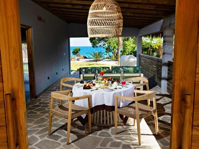 Zastřešená jídelní část s dřevěným nábytkem s výhledem na zahradu a moře.