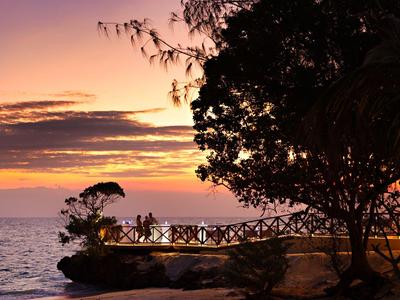 Lidé na vyhlídkové plošině u moře při západu slunce s stromy v popředí.