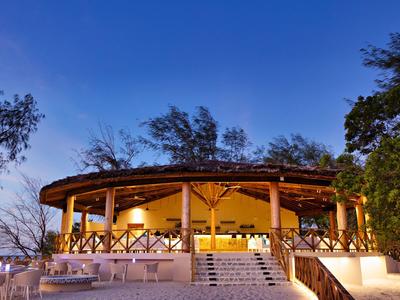 Kruhová restaurace pod širým nebem s dřevěnou konstrukcí a teplým osvětlením v tropickém prostředí za soumraku.