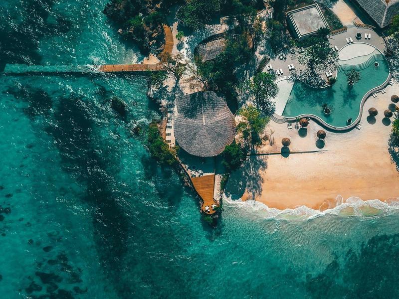 Ptačí pohled na pláž s bazénem a lehátky u tyrkysové vody.