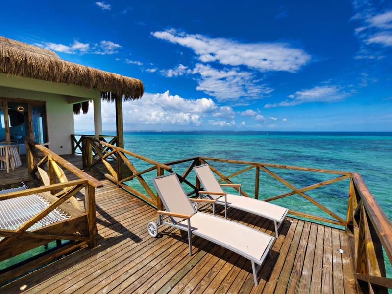 Dřevěná terasa na kůlech s lehátkem nad tyrkysovým mořem pod modrou oblohou.