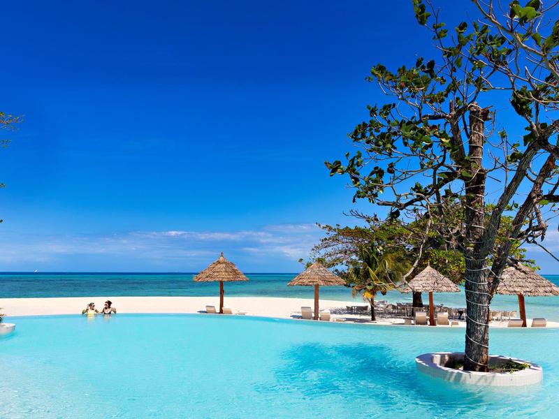 Tyrkysový bazén u bílé písečné pláže s slunečníky a modrou oblohou.