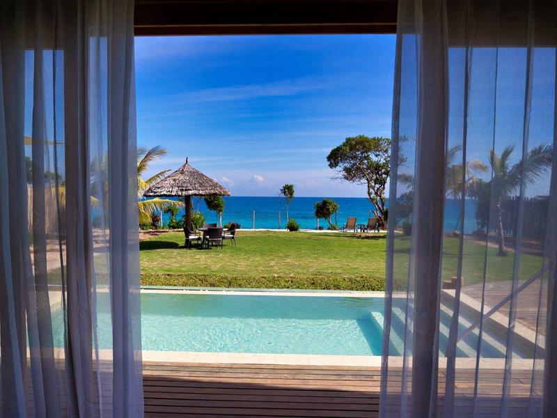 Výhled na bazén a zahradu směrem k moři z místnosti s průhlednými závěsy.