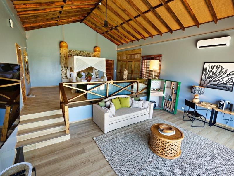 Moderní obývací prostor s bílou pohovkou, ratanovým stolkem a dřevěnou podlahou pod dřevěným stropem.