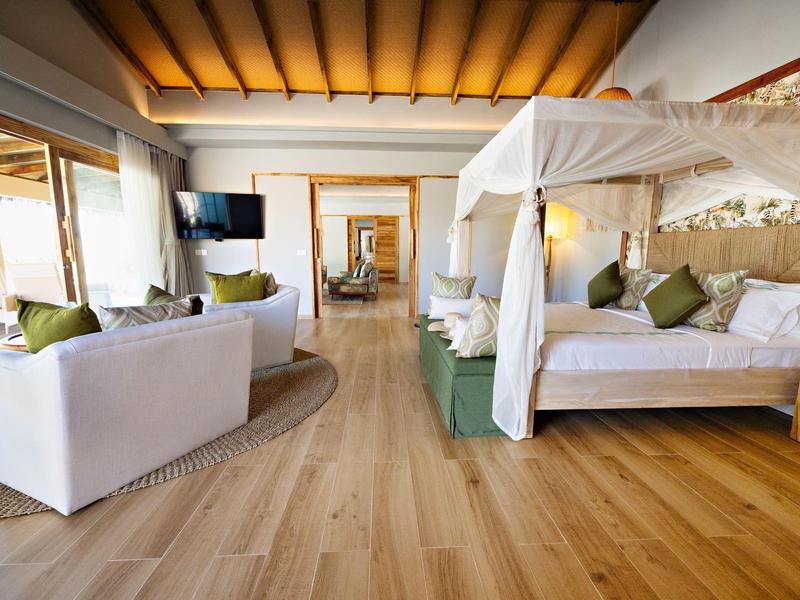 Prostorný hotelový pokoj s baldachýnovou postelí a bílými pohovkami, dostatek přirozeného světla.