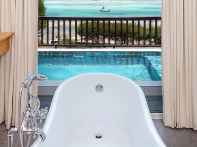 Freestanding bathtub overlooking pool and sea beyond a balcony.