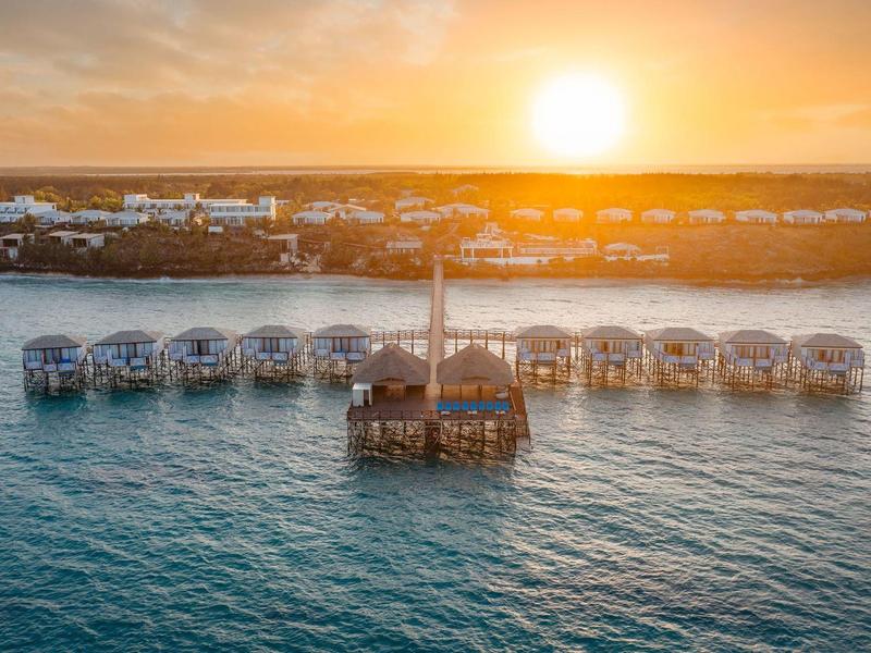 Sunset over water bungalows with pier on clear sea water and coastline in background.
