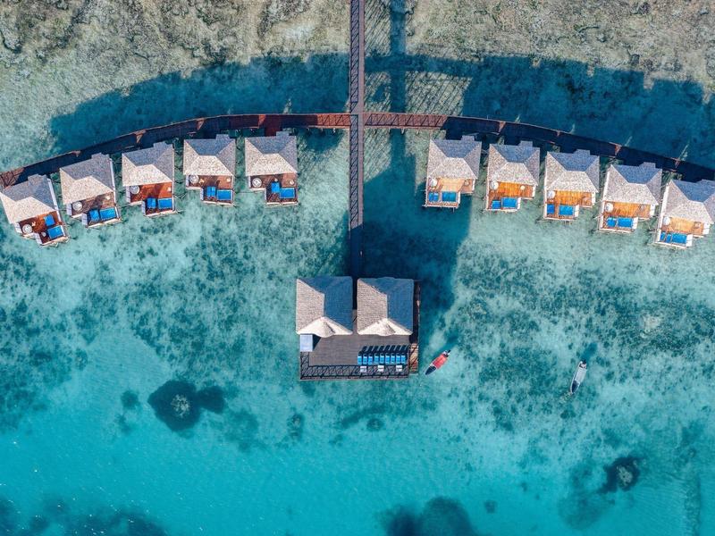 Aerial view of water villas arranged in a semicircle over clear turquoise ocean water.