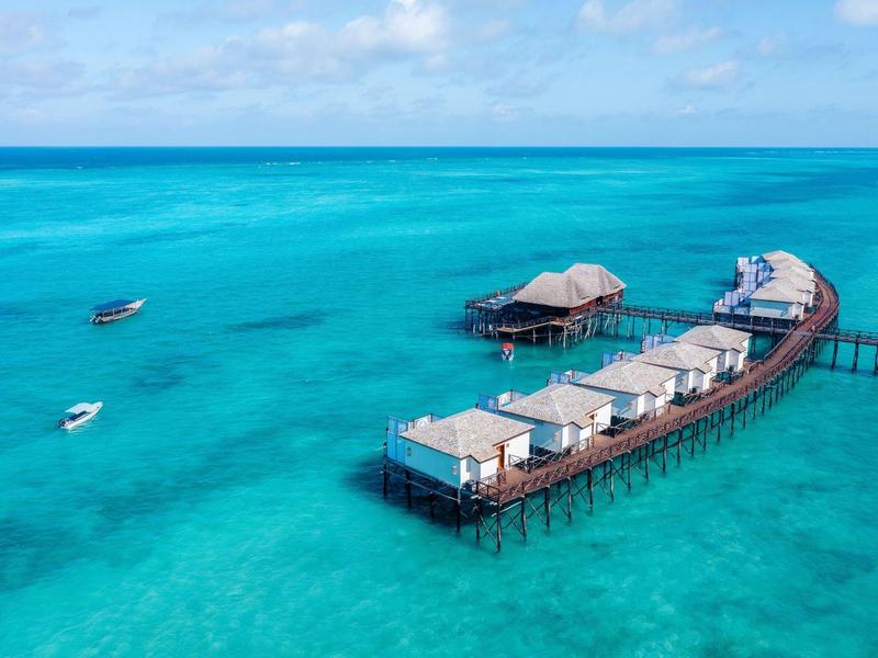 Overwater bungalows on stilts in clear turquoise sea with boats and blue sky.