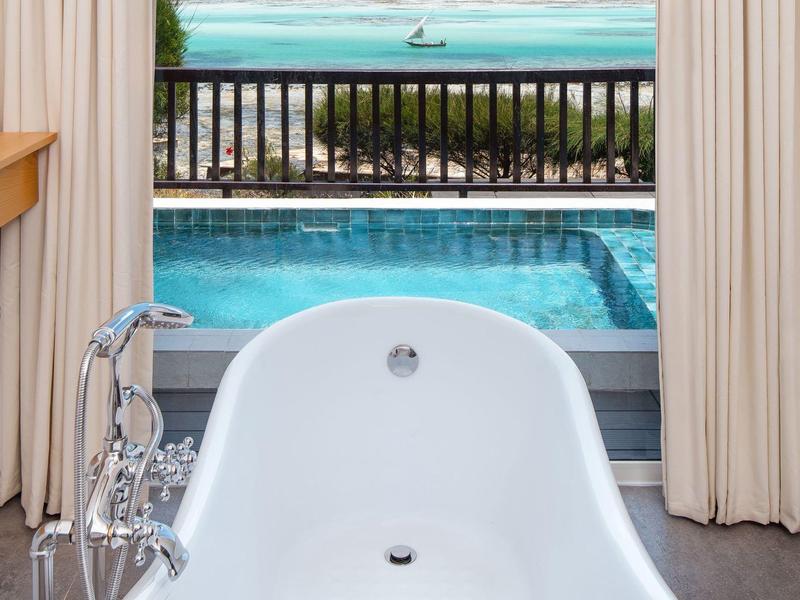 Freestanding bathtub overlooking pool and sea beyond a balcony.