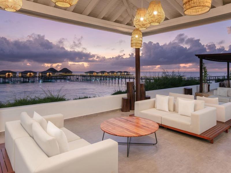 Open lounge area with white sofas overlooking water bungalows at sunset.