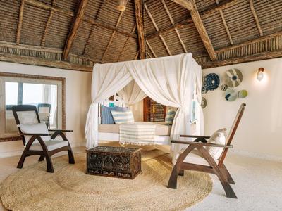 Ein luxuriöses Schlafzimmer mit Himmelbett, zwei Stühlen und einem Teppich unter einem strohgedeckten Dach.