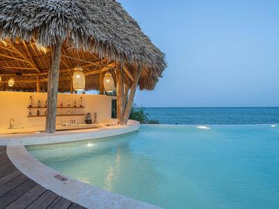 Un bungalow de playa abierto con techo de palma junto a una piscina con vista al mar bajo un cielo despejado.