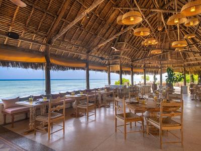 Offenes Strandrestaurant mit Holztischen und -stühlen unter einem Strohdach mit Blick aufs Meer.