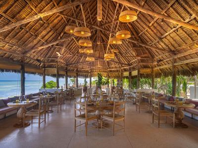 Área de comedor abierta con techo de paja y vista al mar en un entorno tropical.